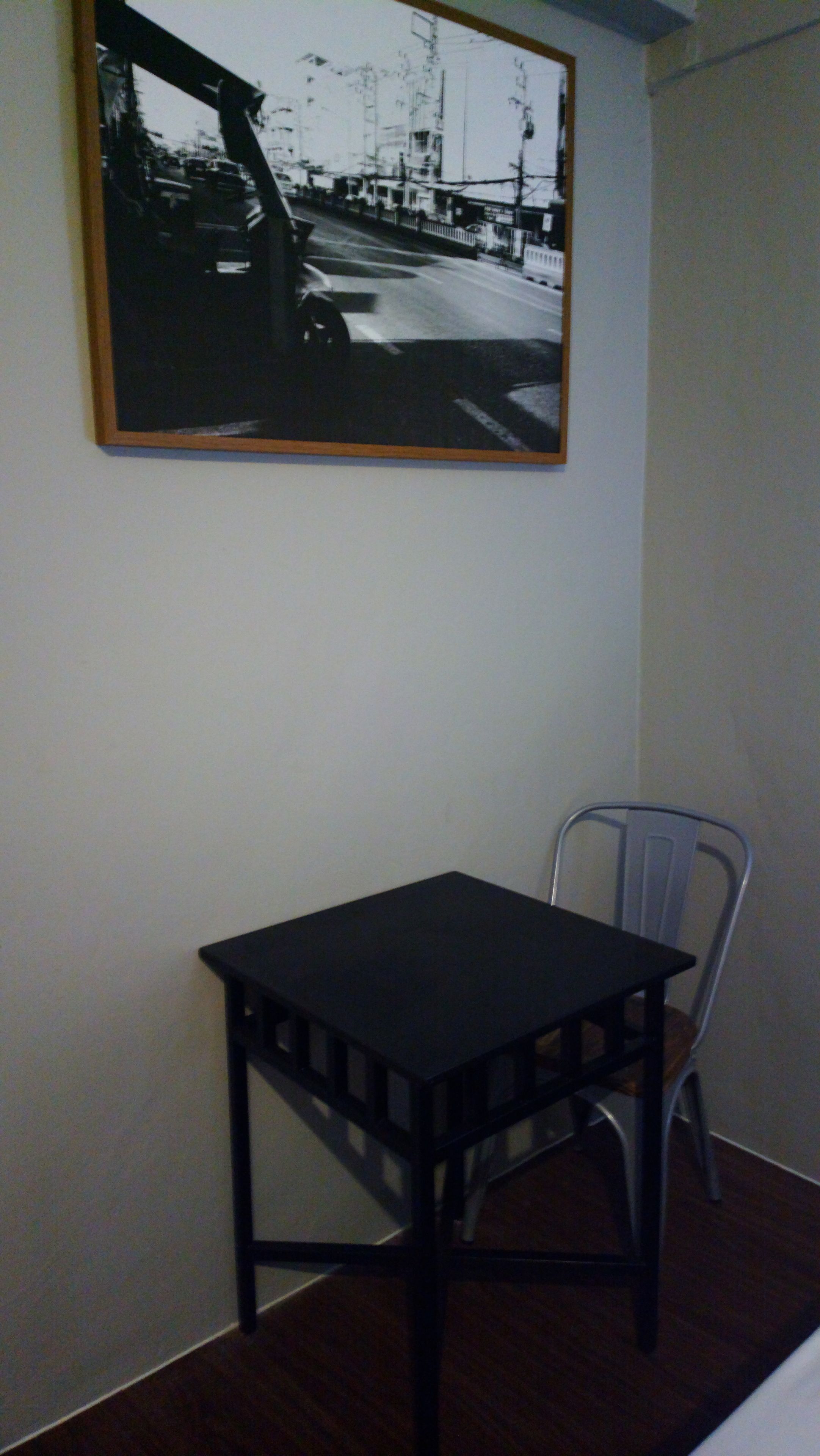 A small room with a black table and a chair, a framed black and white photo on the wall, and a plain white wall.
