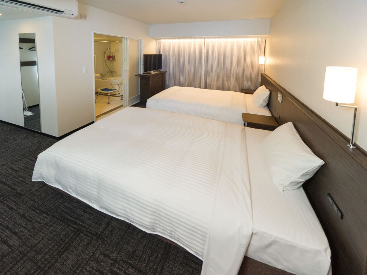Quarto de hotel com uma cama grande, mesa de cabeceira, luminária, TV e banheiro visível por uma porta aberta.