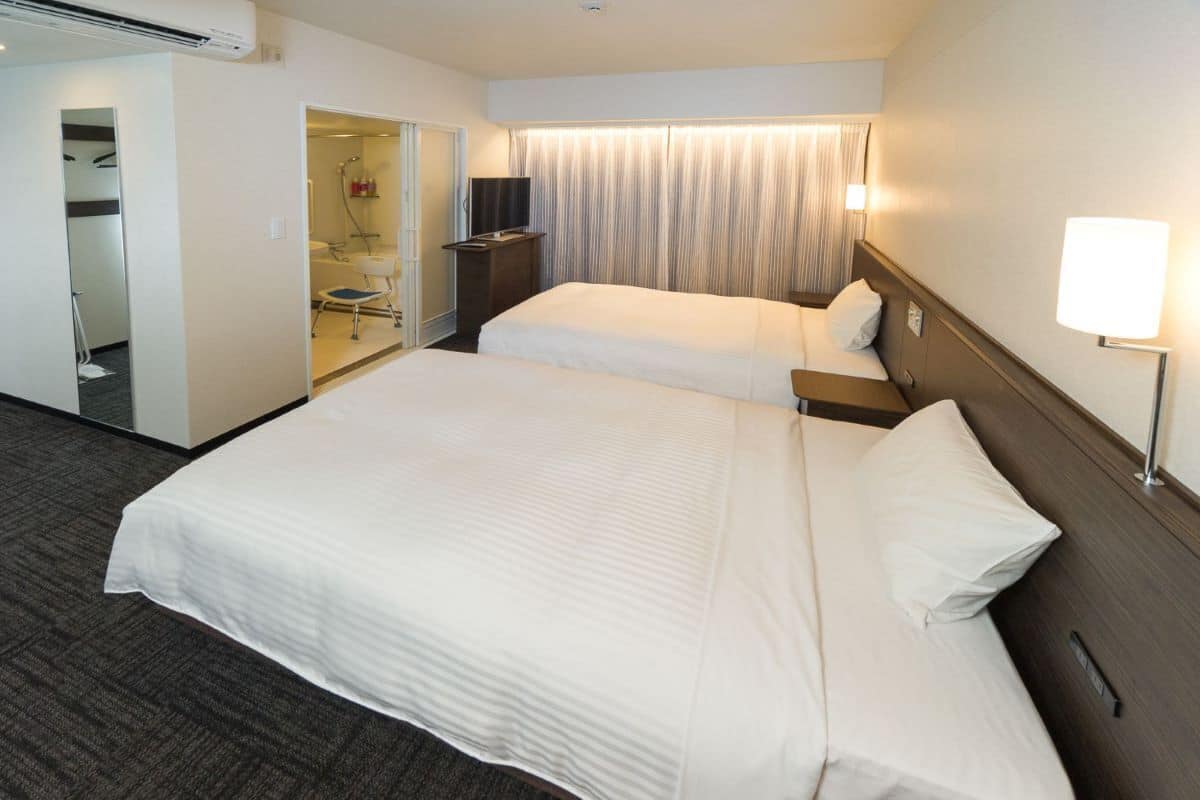 Une chambre d’hôtel avec un grand lit, une table de chevet, une lampe, un téléviseur et une salle de bain visible à travers une porte ouverte.