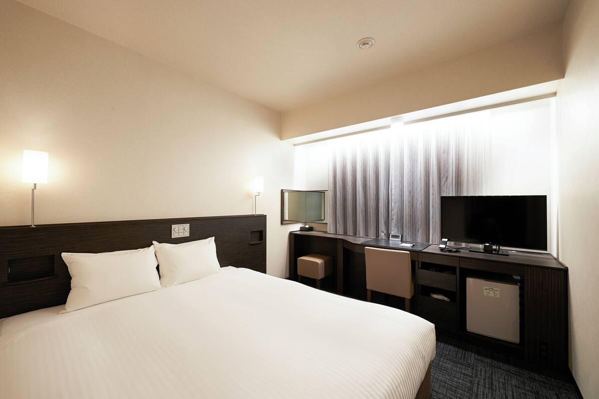 Une chambre d’hôtel avec un grand lit, un bureau avec un ordinateur, une télévision et un petit réfrigérateur.