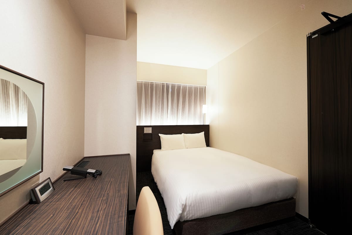 Une chambre d’hôtel avec un lit, un bureau et une chaise, un miroir et une fenêtre avec des rideaux.