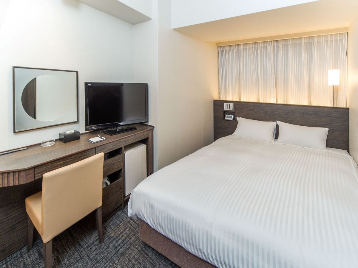 Quarto de hotel com uma cama, uma escrivaninha com televisão, uma cadeira e uma janela com cortinas.