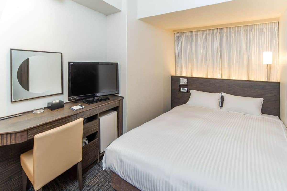 Une chambre d’hôtel avec un lit, un bureau avec une télévision, une chaise et une fenêtre avec des rideaux.