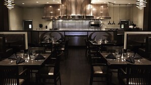 A modern restaurant with dark wood furniture, a central kitchen area, and pendant lighting.