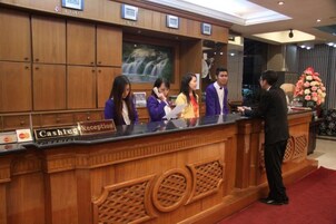 A hotel reception desk with a wooden finish, a reception area, and a large floral arrangement in the background.