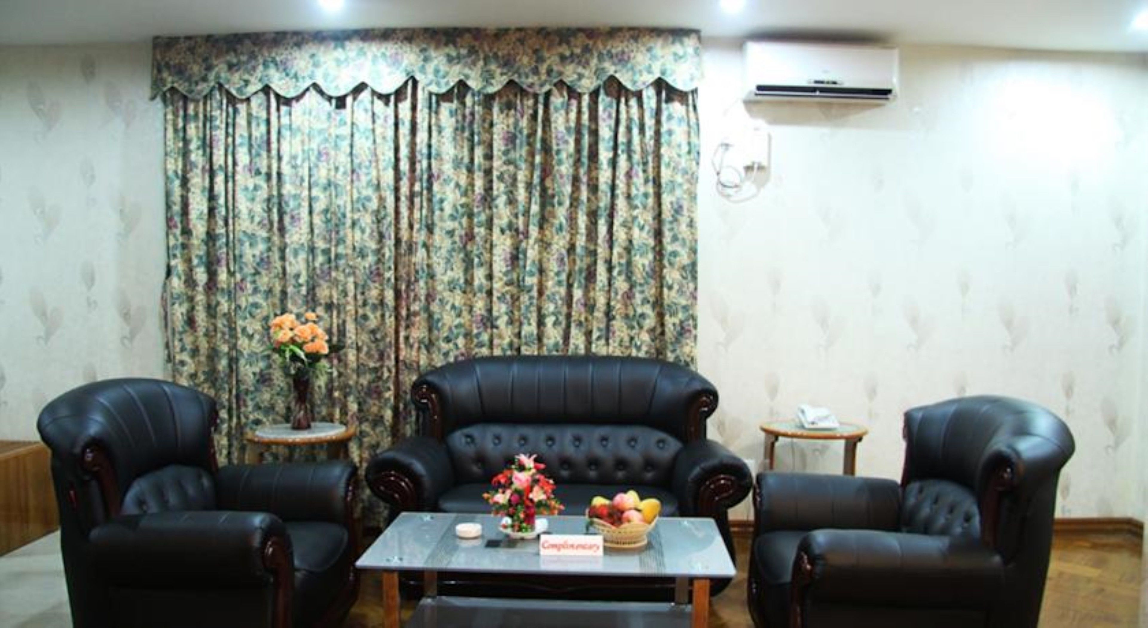 Una habitación con un sofá de cuero negro, dos sillones, una mesita con un florero y una cortina estampada.