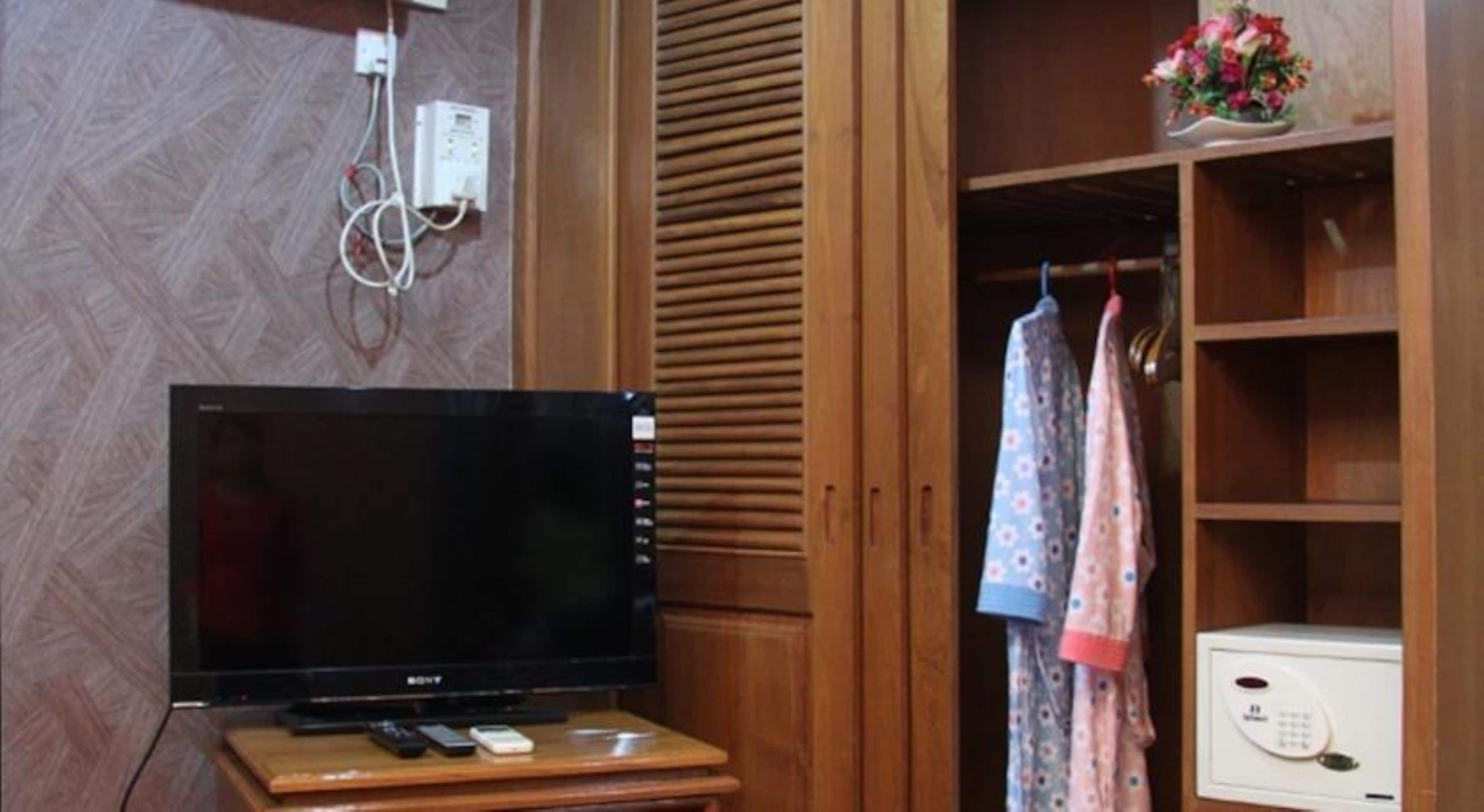 A room with a wooden wardrobe, a television on a wooden stand, and a small table with a remote control and a mobile phone.