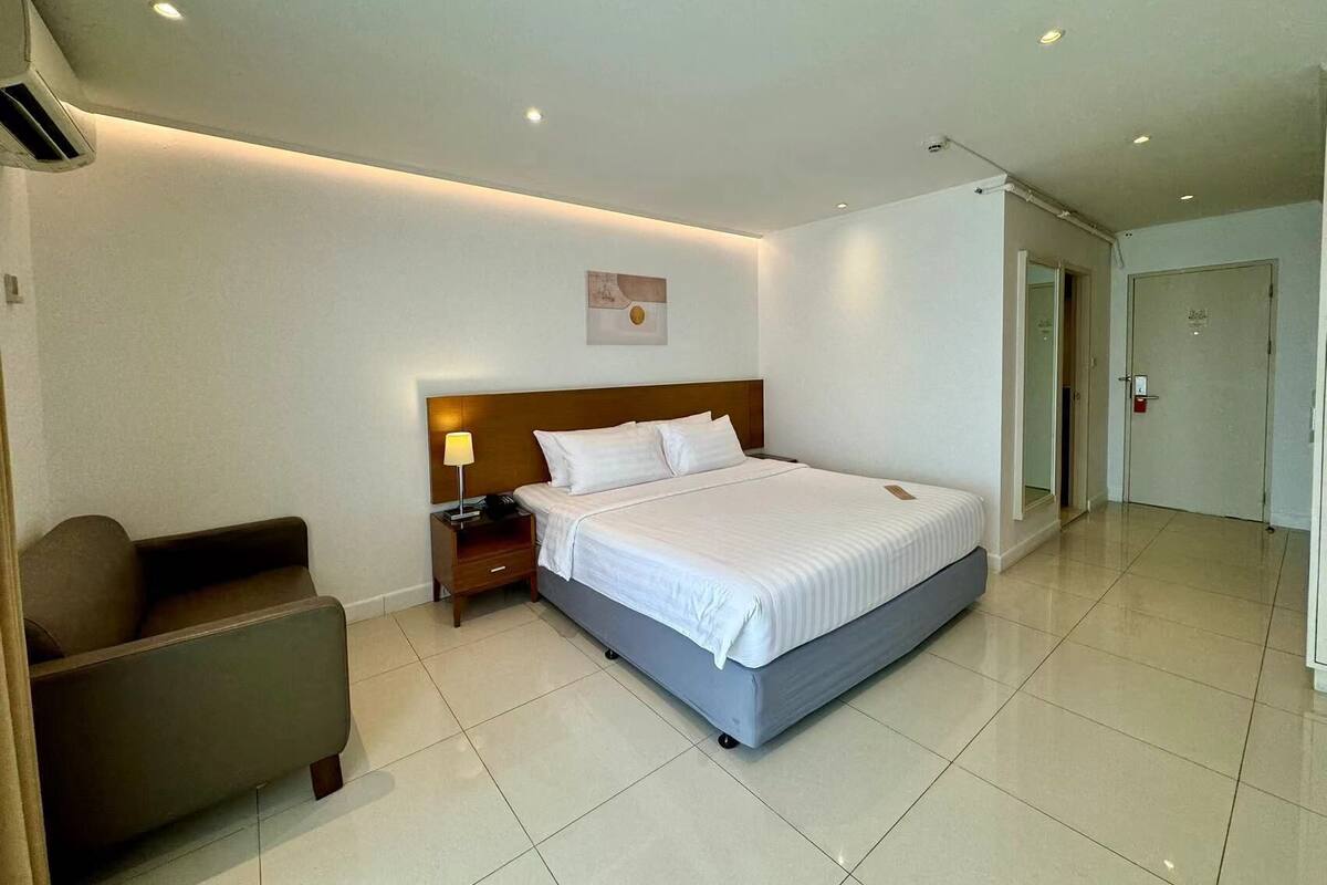 Una habitación de hotel con una cama grande, una mesita de noche con lámpara, una silla y una puerta.