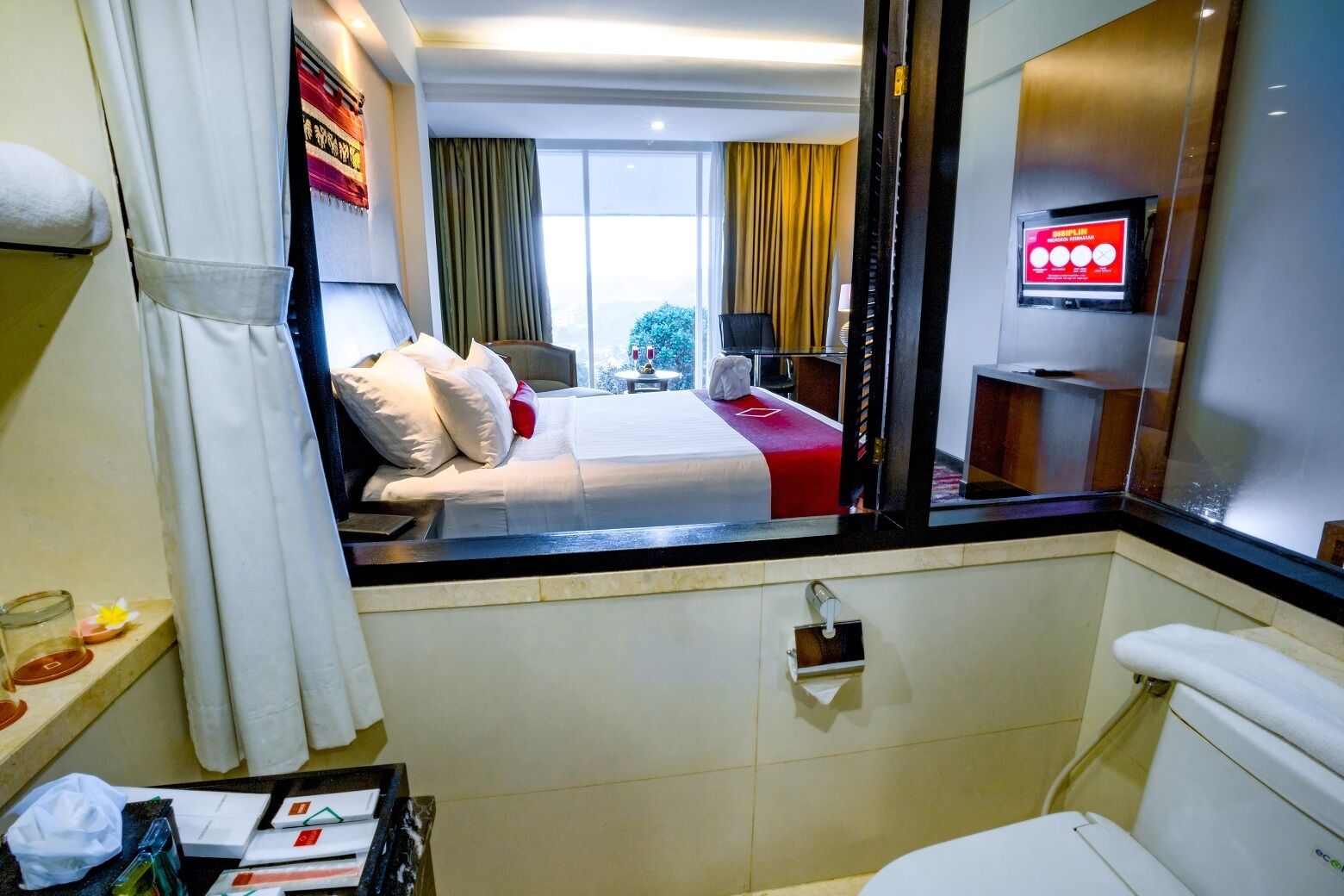 Quarto de hotel com cama, uma pequena mesa com abajur, janela com cortinas e um espelho.