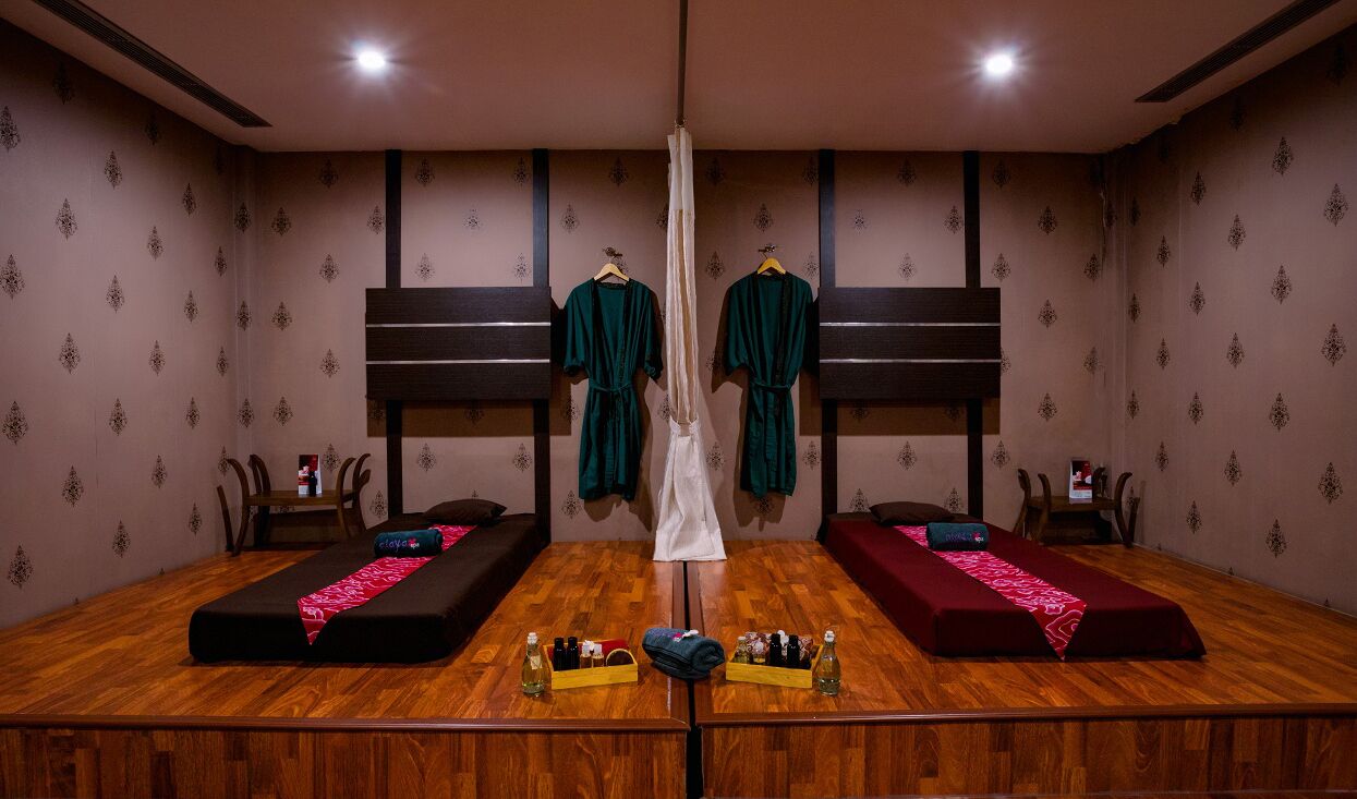 A room with two beds, a wooden floor, and a mirror. There are two wooden headboards, a white curtain, and a small table with bottles and a tissue box.