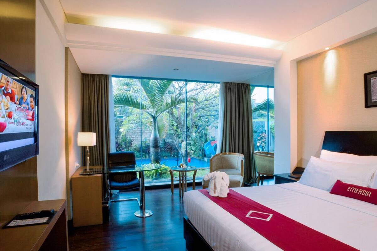 Une chambre d’hôtel avec un grand lit, une télévision, un bureau, une chaise et une vue sur la verdure grâce à de grandes fenêtres.