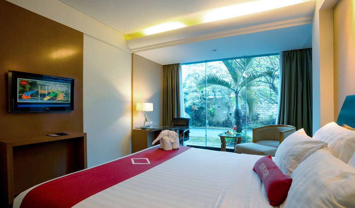 Habitación de hotel con una cama grande, un televisor en la pared, un escritorio con lámpara y vistas a palmeras a través de la ventana.