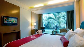 Habitación de hotel con una cama grande, un televisor en la pared, un escritorio con lámpara y vistas a palmeras a través de la ventana.