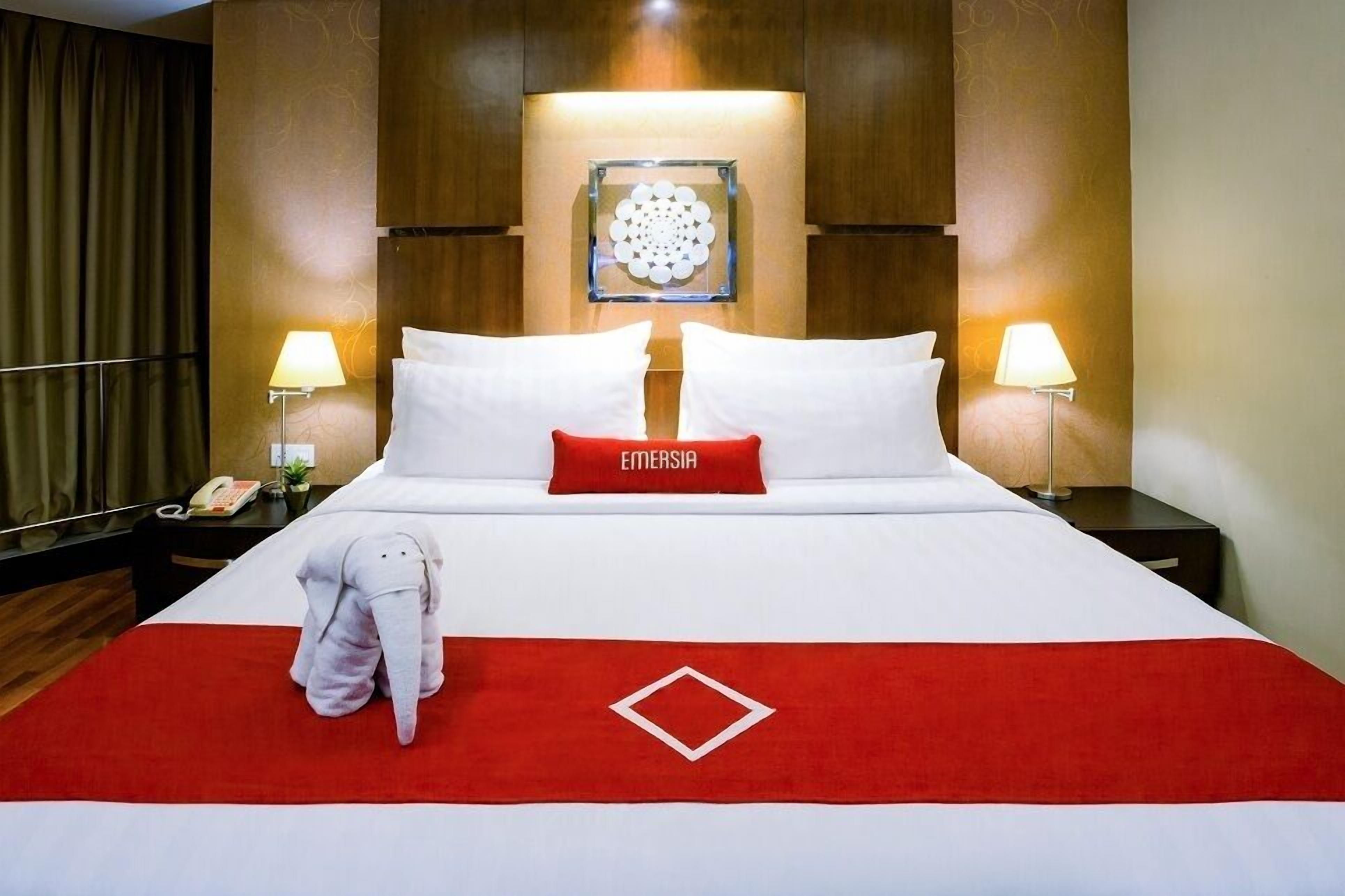 A hotel room with a large bed, two bedside lamps, a decorative wall piece, and a red pillow with the word "EMERSIA".