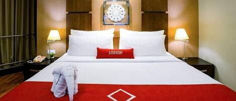 Habitación de hotel con una cama grande, dos lámparas de noche, un adorno en la pared y una almohada roja con la palabra “EMERSIA”.