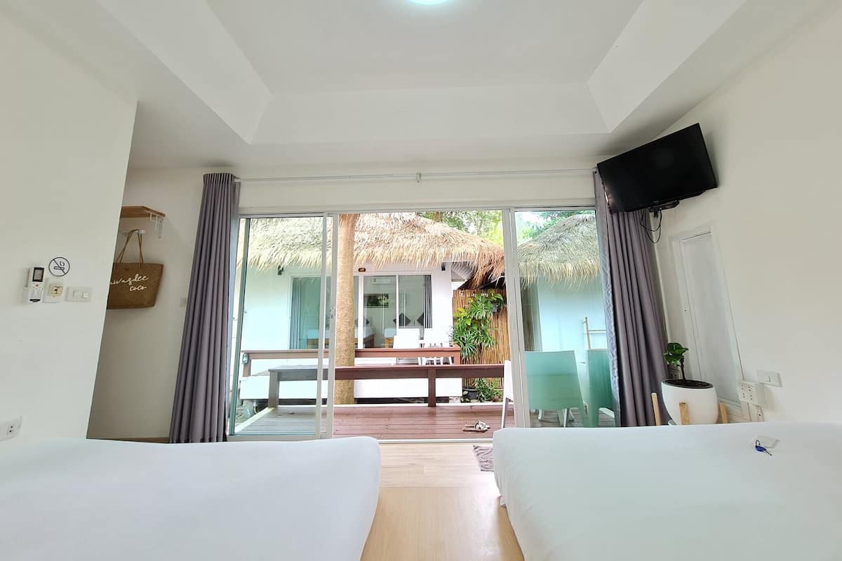 A hotel room with two beds, a TV, a balcony with a thatched roof, and a view of the outdoor area.