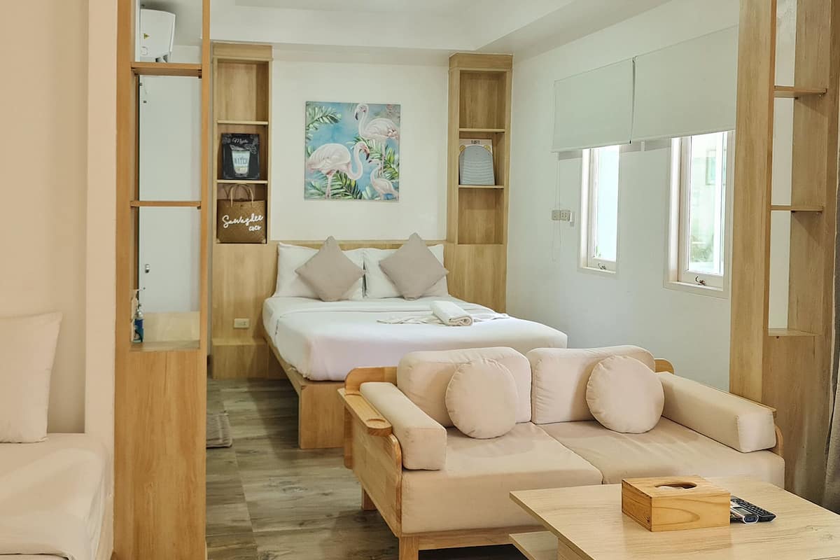 A compact hotel room with a bed, a sofa, a wooden side table, and a small wooden shelf with personal items.