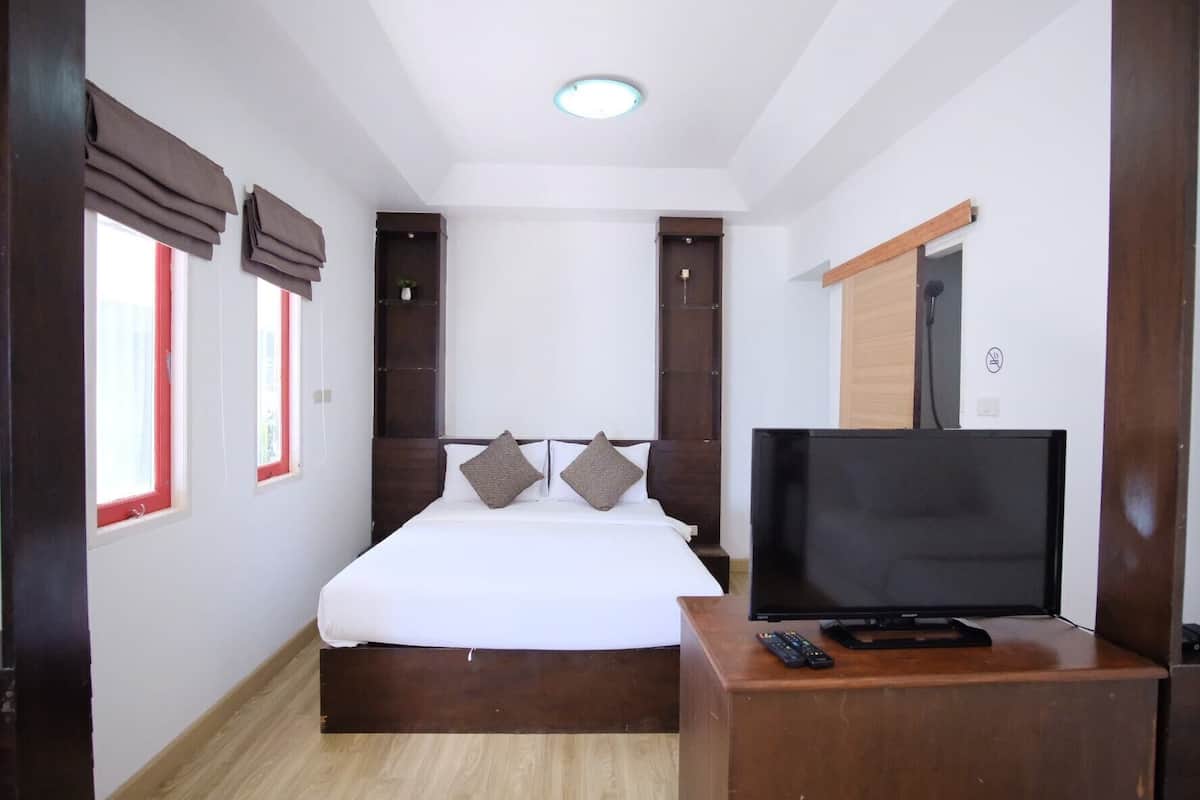 A bedroom with a bed, a TV on a wooden stand, a door, and windows with red frames.