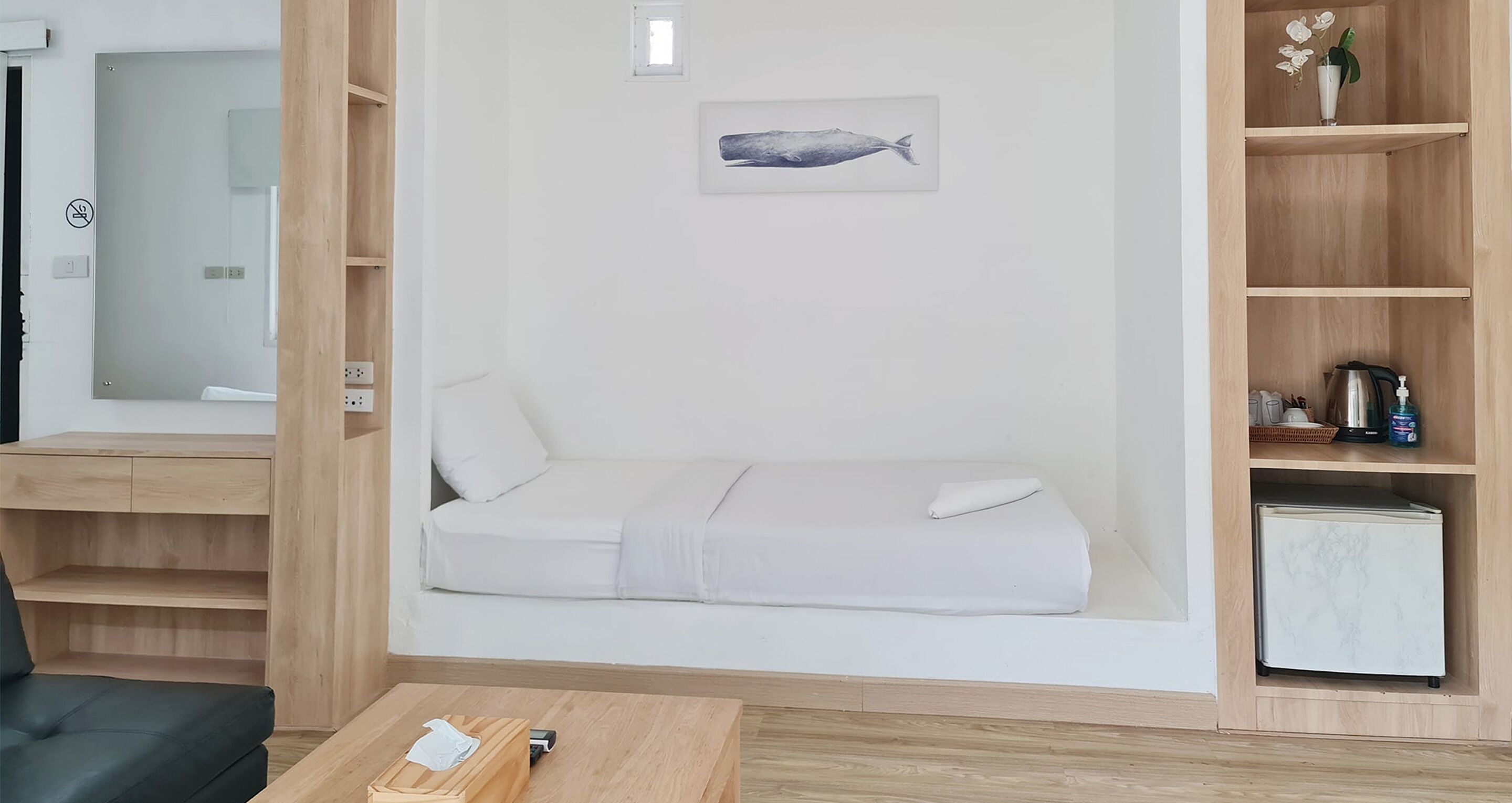 Et minimalistisk rum med en hvid sofa, en lille seng, en træhylde med et maleri af en fisk og et hvidt køleskab.