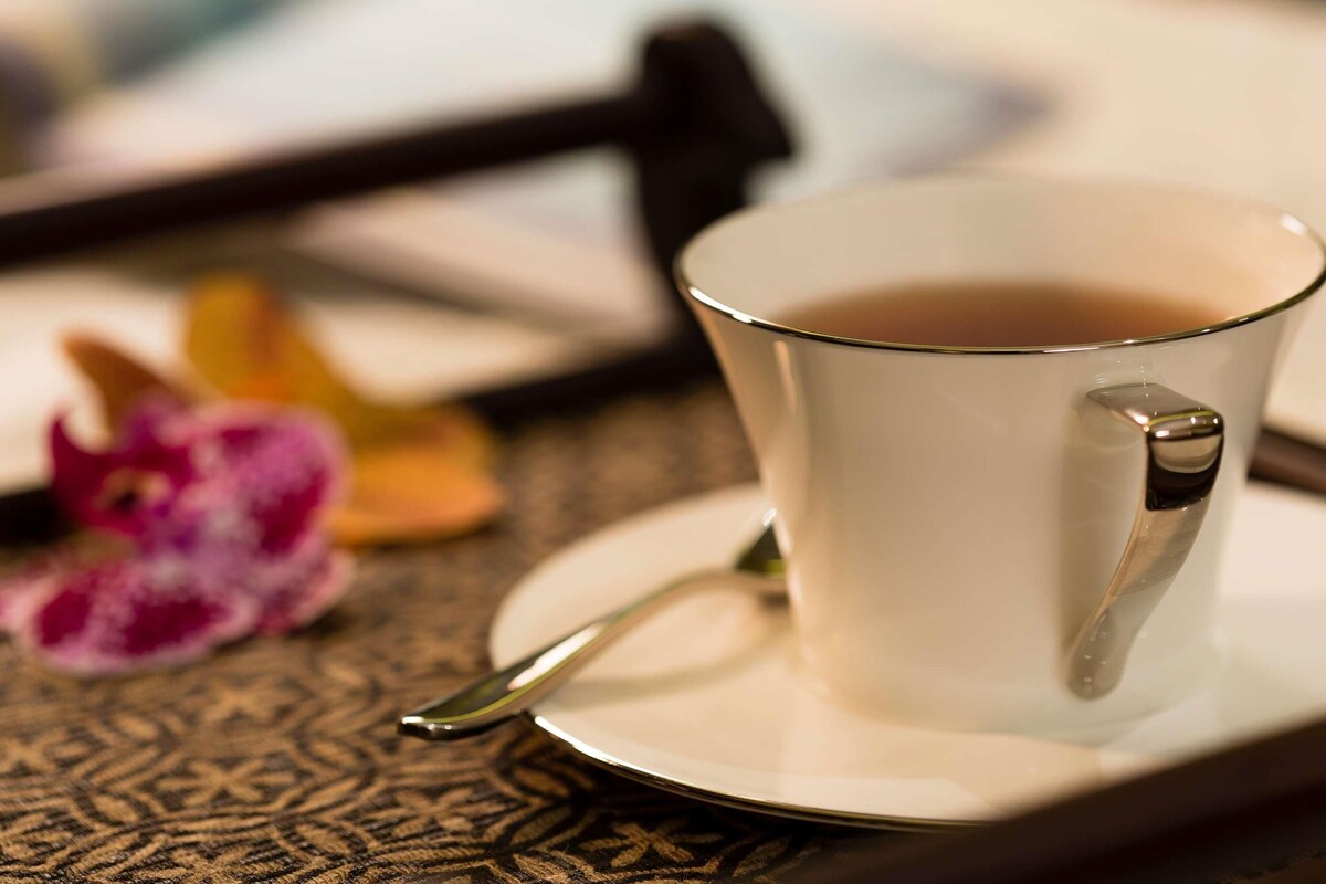 A cup of tea on a saucer with a spoon, a flower, and a blurred background.