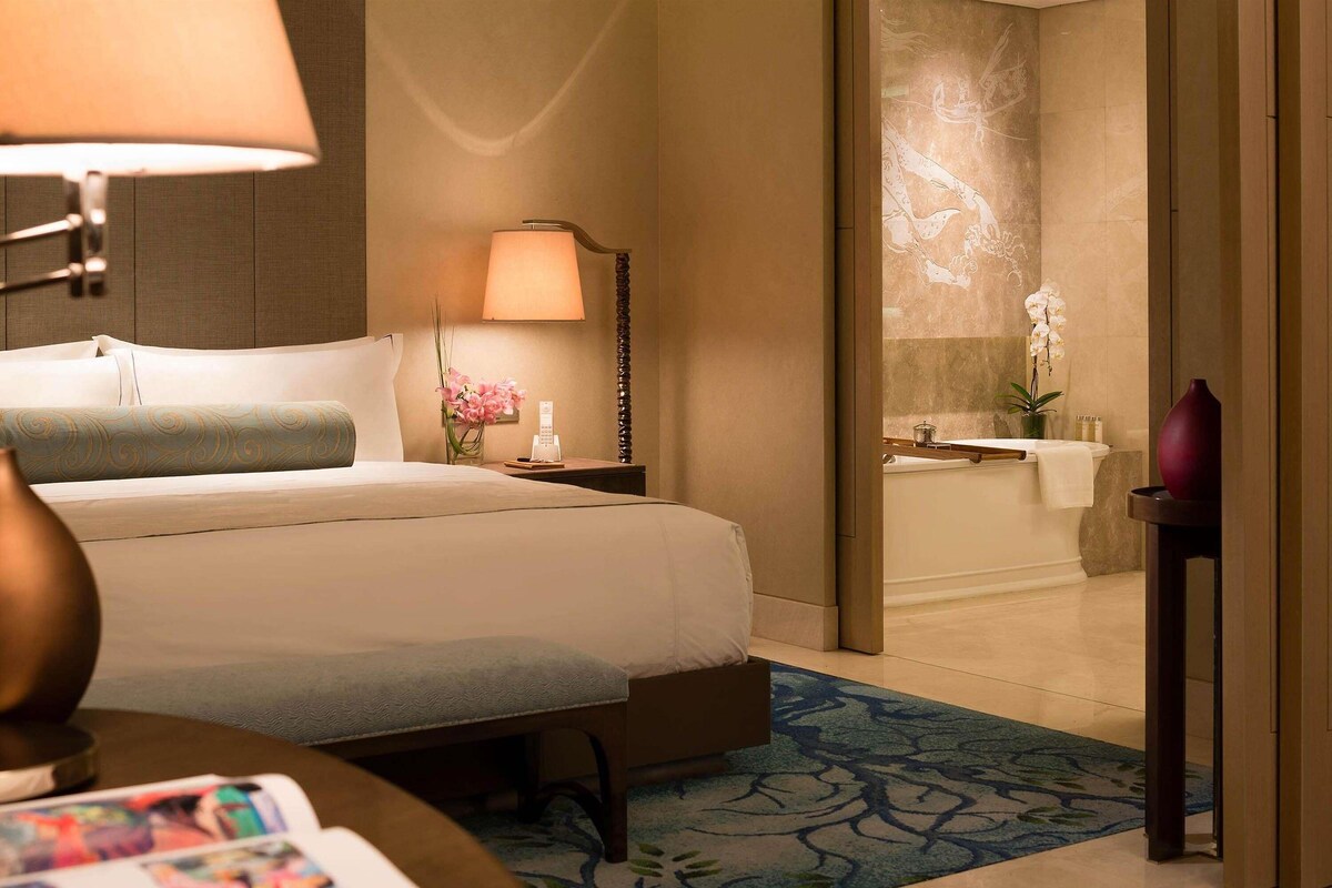 A hotel room with a bed, bedside table, lamp, and a view of the bathroom.