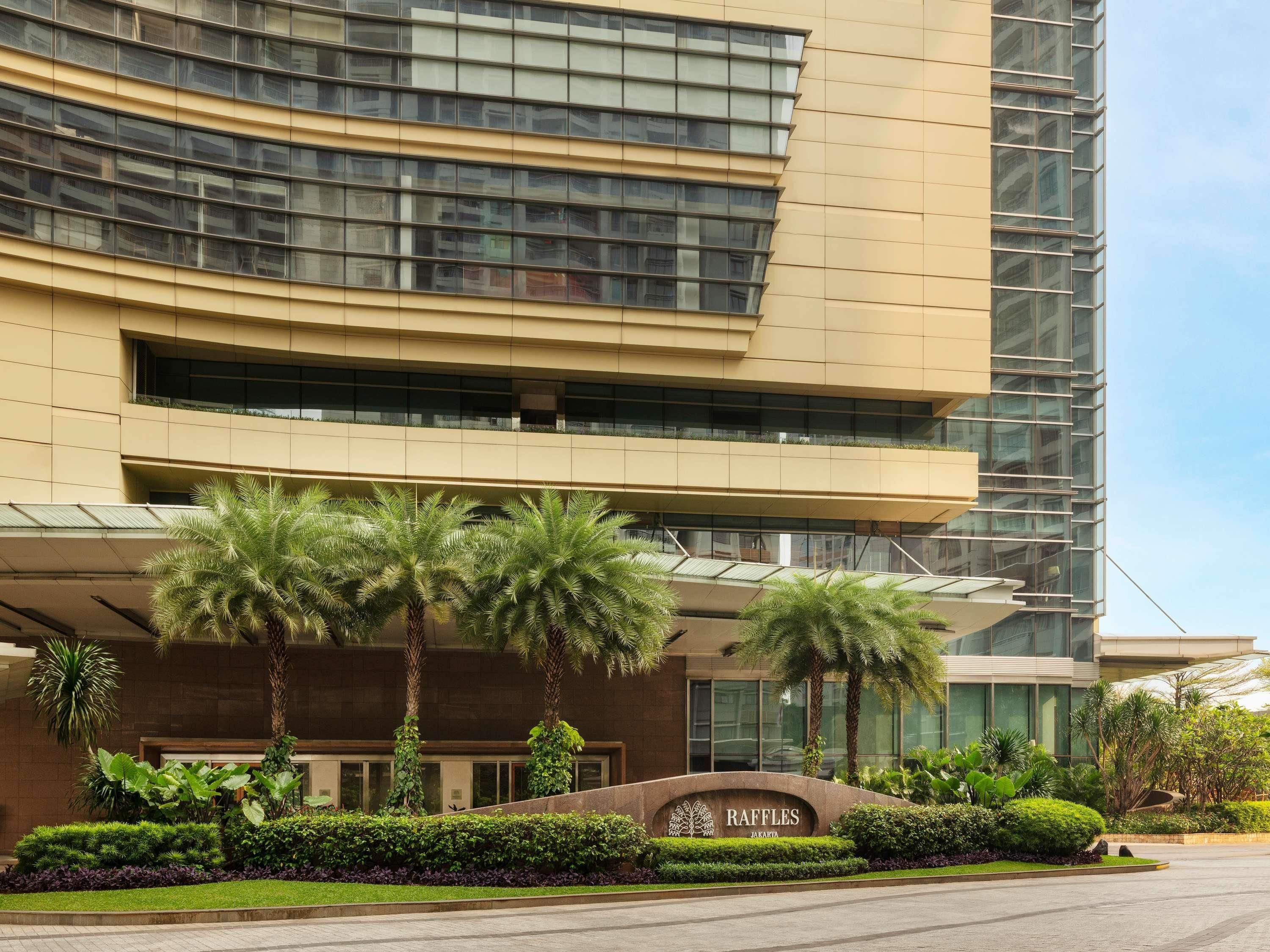 Un edificio moderno con fachada curva, una entrada ajardinada con palmeras y un letrero que dice "RAFFLES JAKARTA".