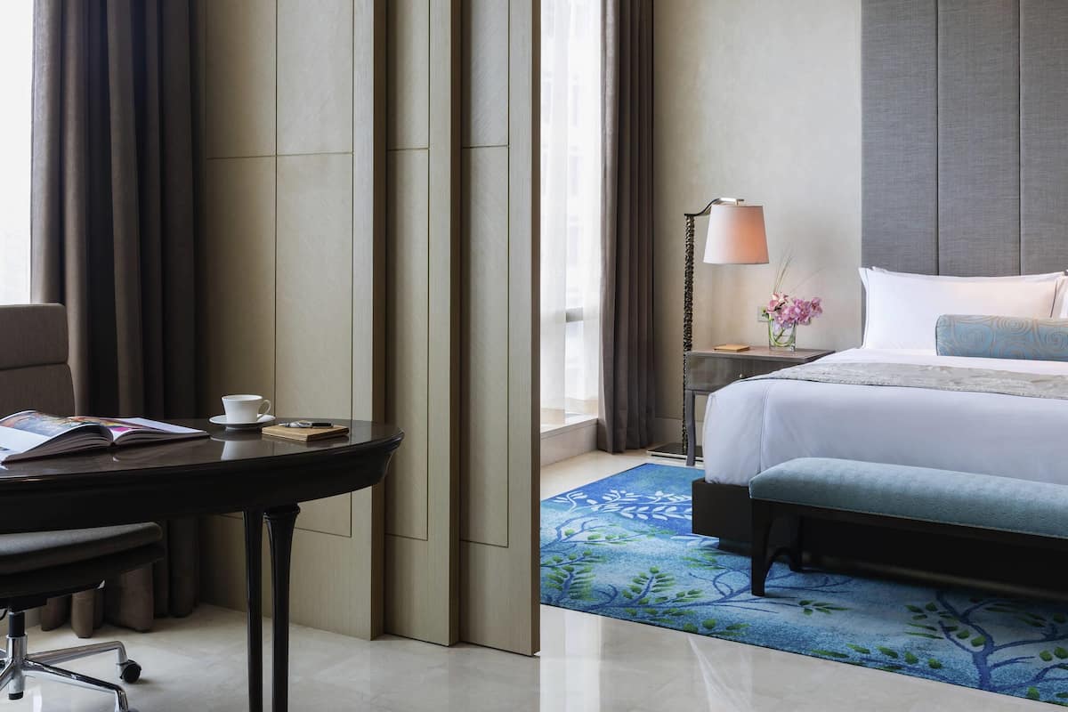 A hotel room with a bed, bedside table, lamp, and a desk with a chair.