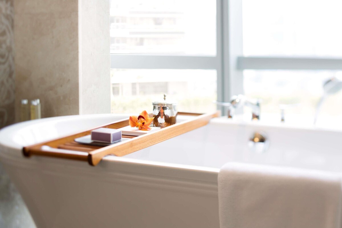 A modern bathroom with a freestanding bathtub, a wooden tray with toiletries, and a view of buildings outside.