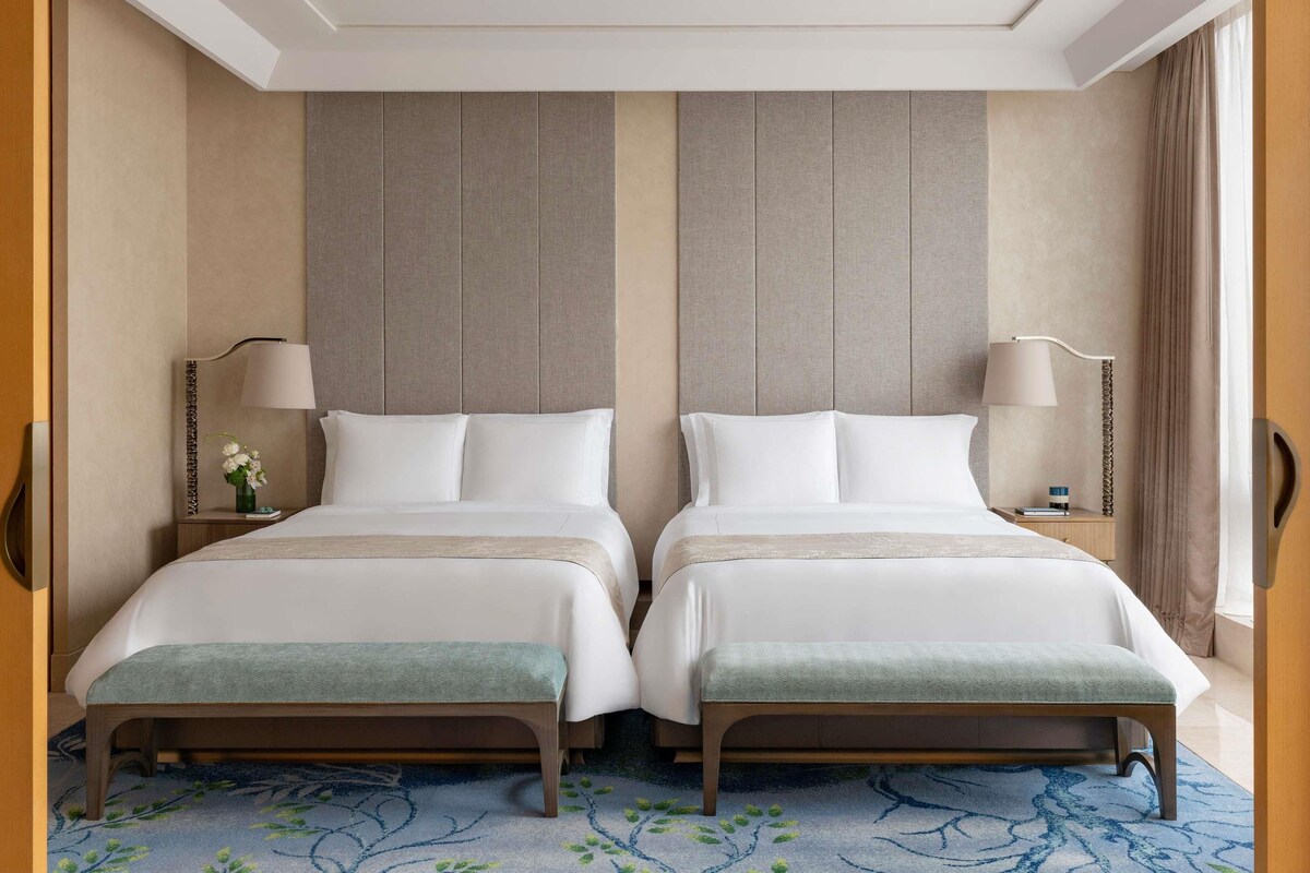 Una habitación de hotel con dos camas, un banco, una mesita de noche con lámpara y un jarrón decorativo.