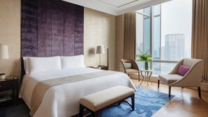 A modern hotel room with a large bed, a bench, a chair, a small table with a lamp, and a view of the city through large windows. - Raffles Jakarta (Jakarta)