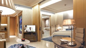 A modern hotel room with a flat-screen TV, a bed, a desk with a lamp, a chair, and a round table with a vase. - Raffles Jakarta (Jakarta)