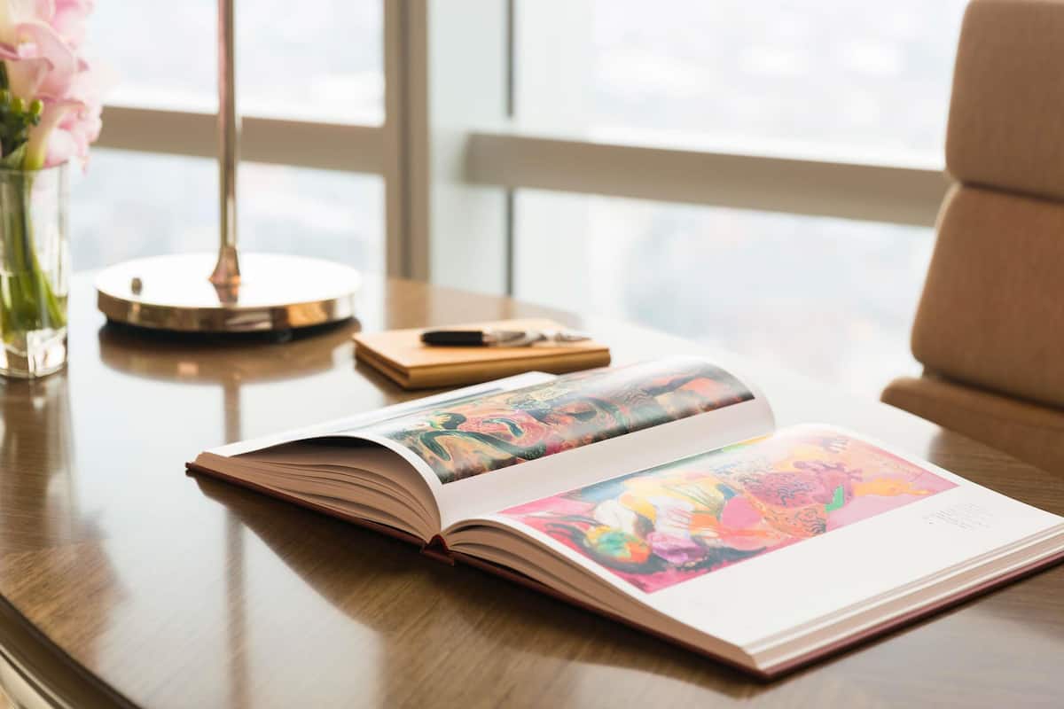 An open book with a colorful illustration on a wooden table, a vase with pink flowers, and a lamp.