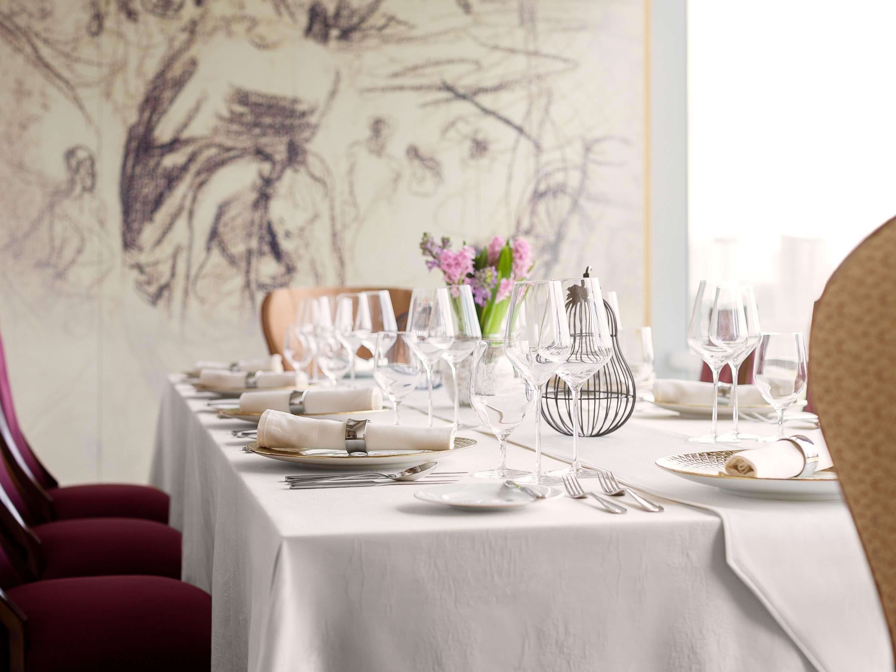 A dining table set with white tablecloth, silverware, and wine glasses, with a floral centerpiece and a wall painting in the background.