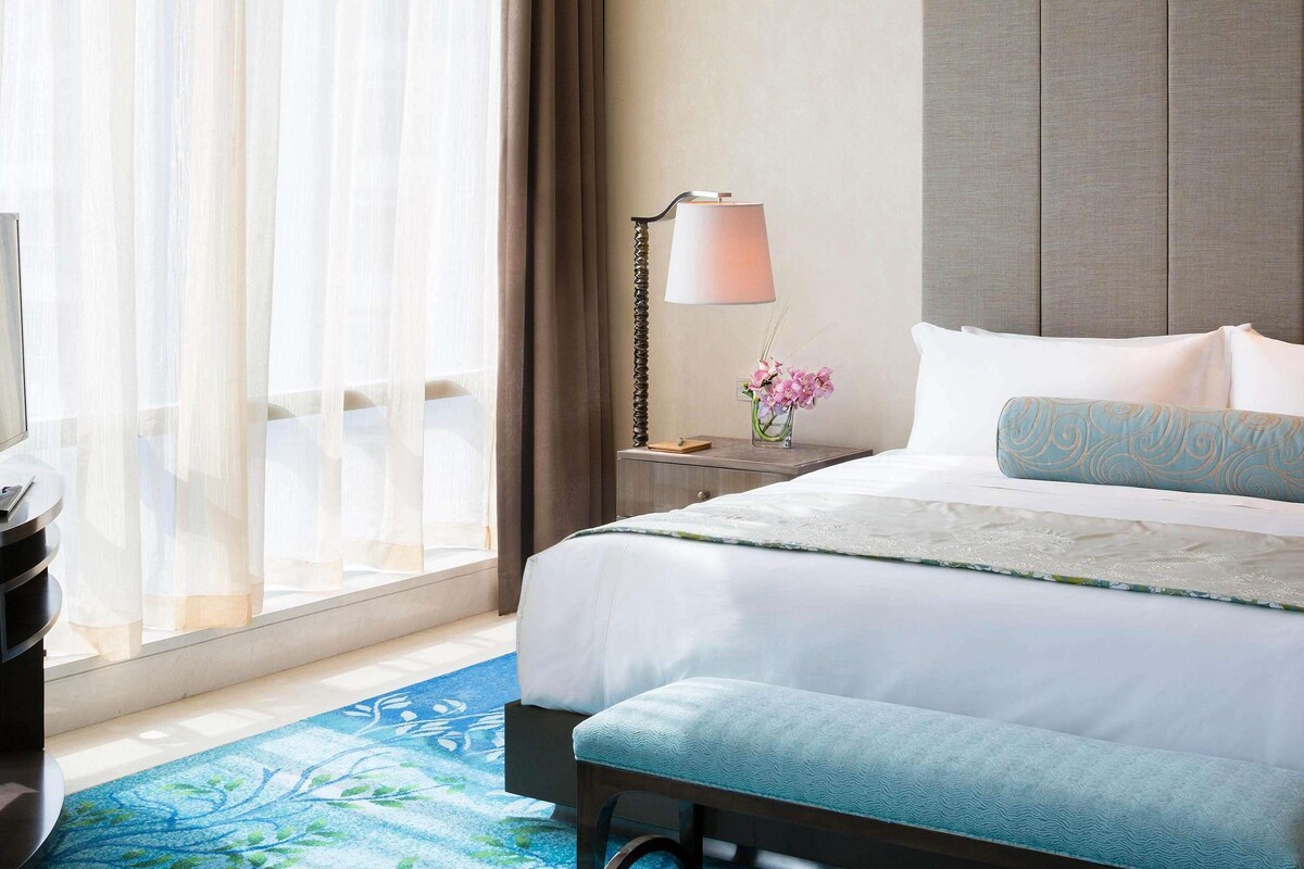 Una habitación de hotel con una cama, una mesita de noche con una lámpara, un jarrón de flores y una alfombra azul estampada.