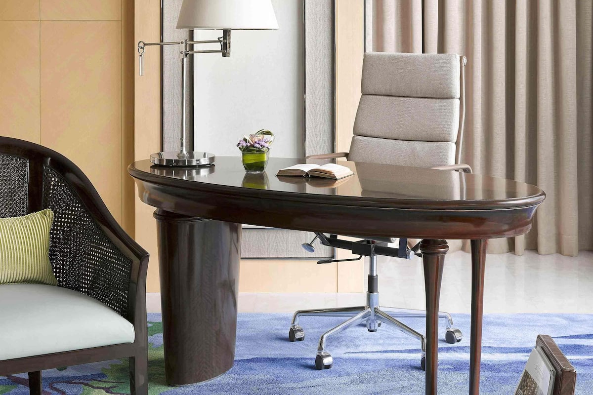 A modern hotel room with a glass-top desk, a comfortable chair, a lamp, and a vase with flowers.