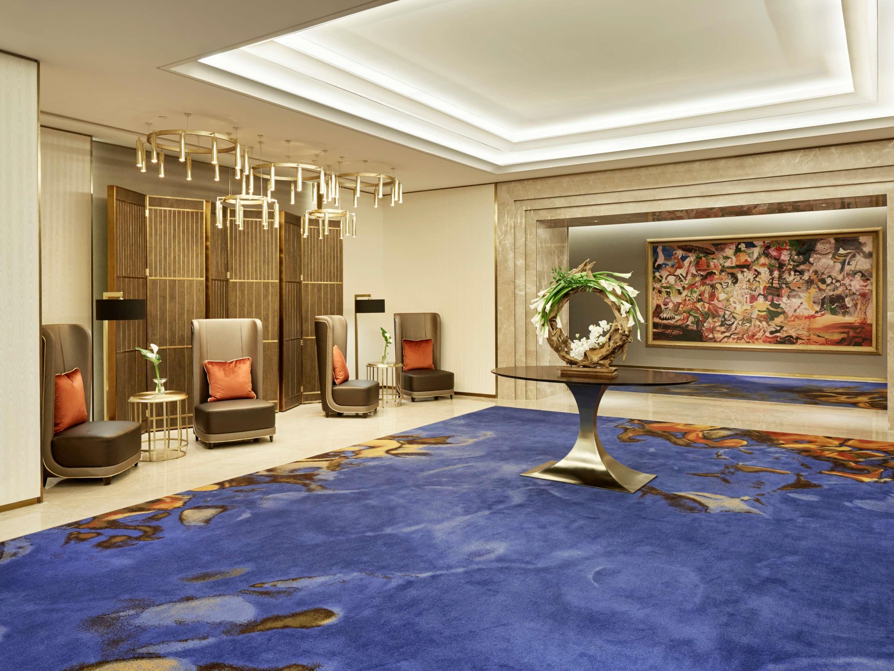 Un moderno vestíbulo de hotel con alfombra azul, zona de estar con sofás y sillones, una mesa redonda con un centro decorativo y un cuadro grande en la pared.