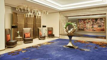 Un moderno vestíbulo de hotel con alfombra azul, zona de estar con sofás y sillones, una mesa redonda con un centro decorativo y un cuadro grande en la pared.
