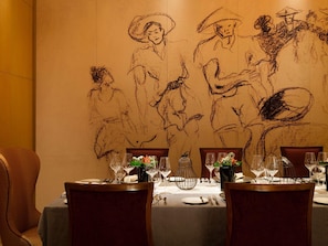 Una mesa de comedor dispuesta con copas de vino, platos y cubiertos, con un gran mural en la pared que representa a personas con vestimenta histórica.
