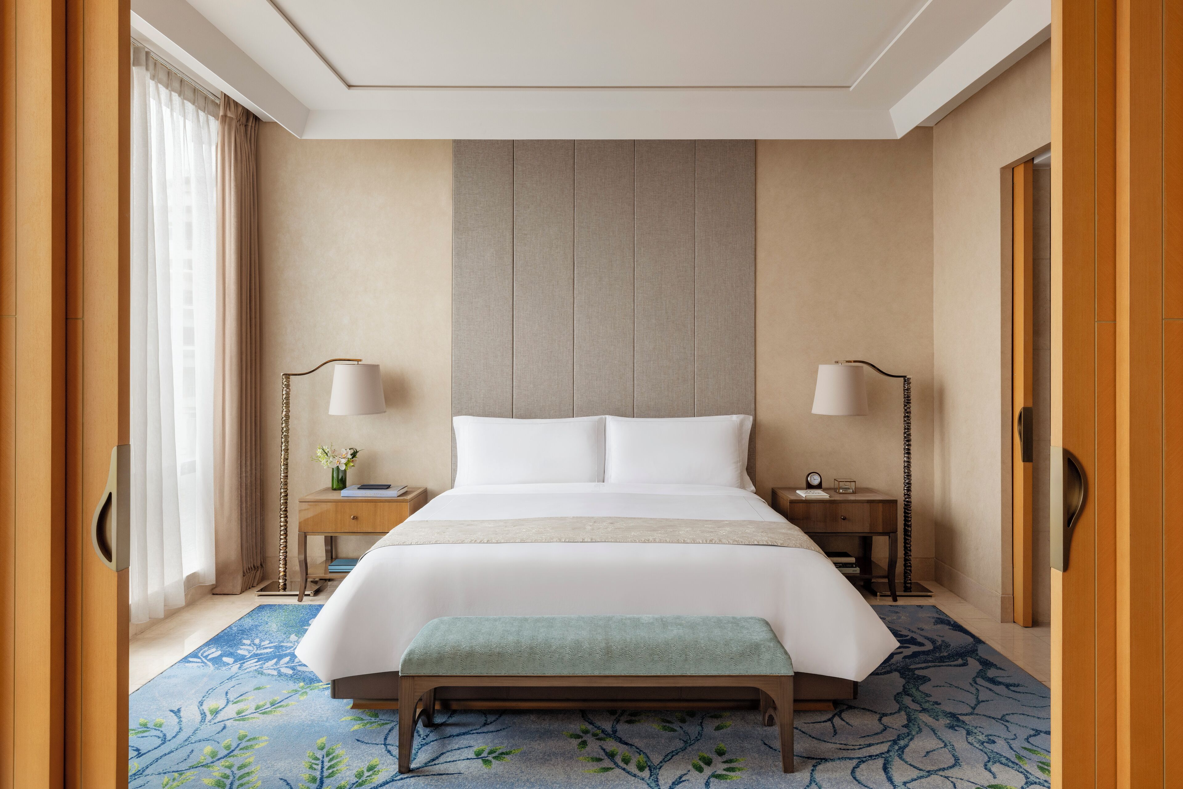 A hotel room with a large bed, two bedside tables, a bench, and a view of the outside through a window with curtains.