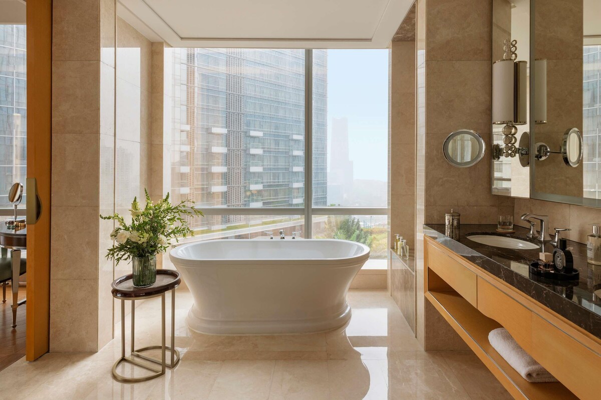 A modern bathroom with a freestanding bathtub, a large mirror, and a view of tall buildings through large windows.