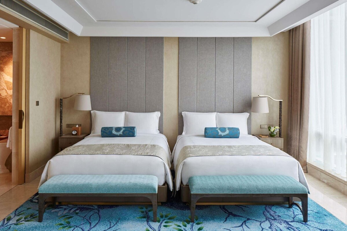 A hotel room with two beds, a large window, and a blue patterned carpet.
