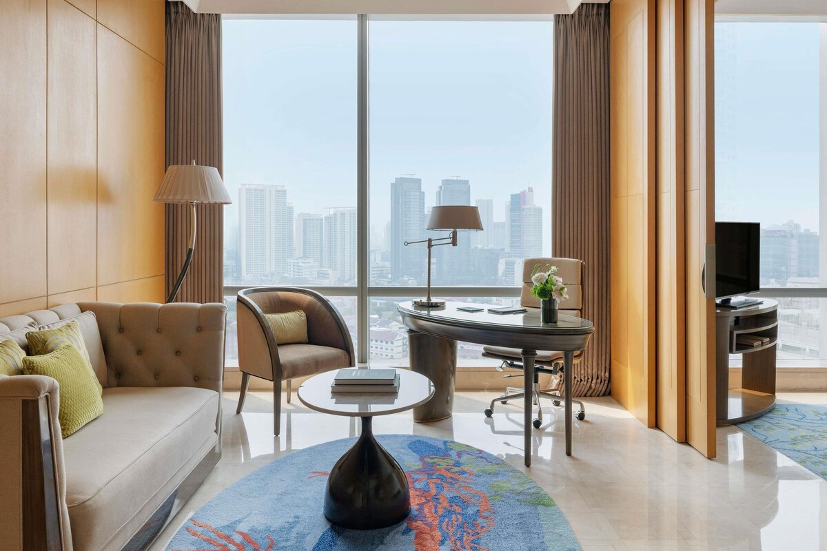 A modern hotel room with a sofa, armchair, desk, and a large window with a city view.