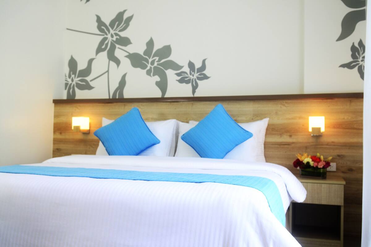 A hotel room with a wooden headboard, two blue pillows, a white bedspread, and a nightstand with a vase of flowers.