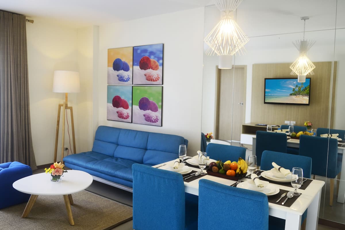 A modern hotel room with a blue sofa, a dining table set for two, and a fruit basket.