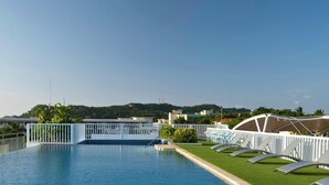 A swimming pool with a white fence, lounge chairs, and a view of buildings and greenery in the background.