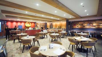 Un restaurant avec des tables et des chaises rondes, un comptoir où se trouve un employé, et une bibliothèque en bois à l’arrière-plan.