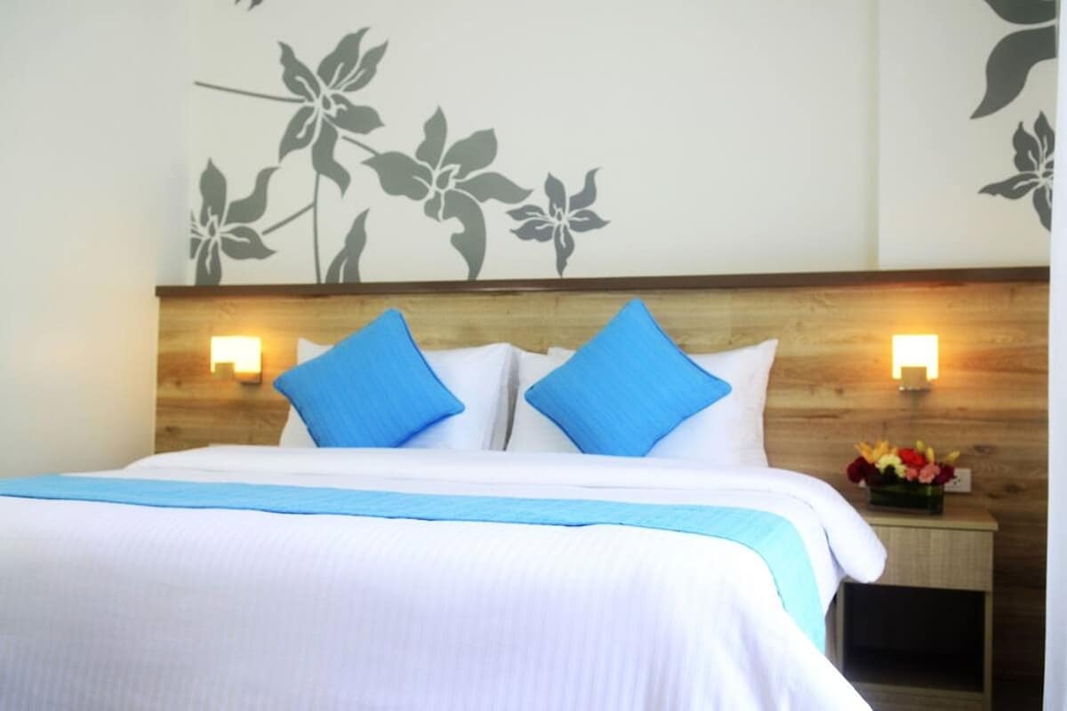 Habitación de hotel con cama, dos almohadas azules, cabecera de madera y luces fijadas a la pared.