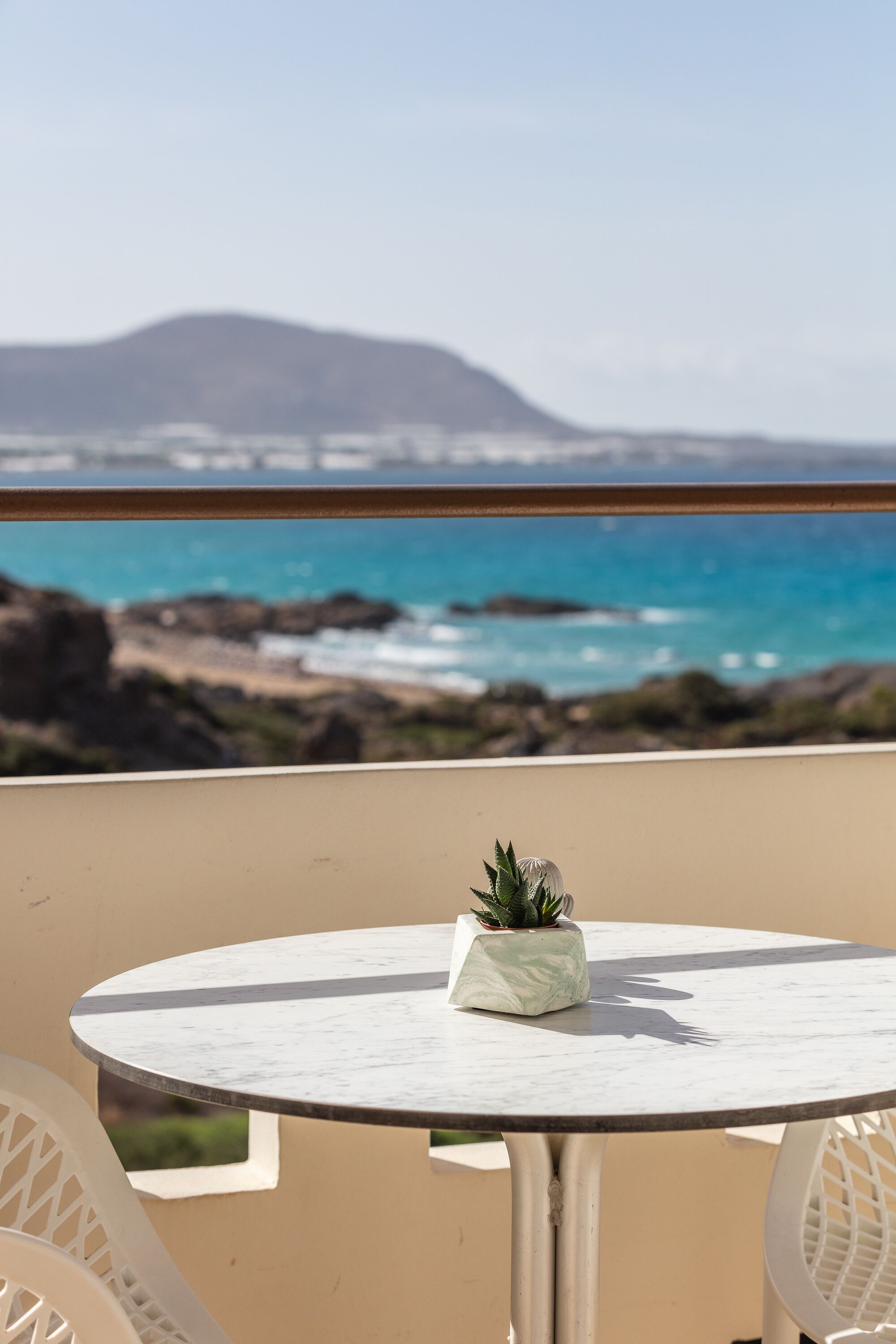 A round table with a potted plant, overlooking a coastal view with rocks and a distant mountain.