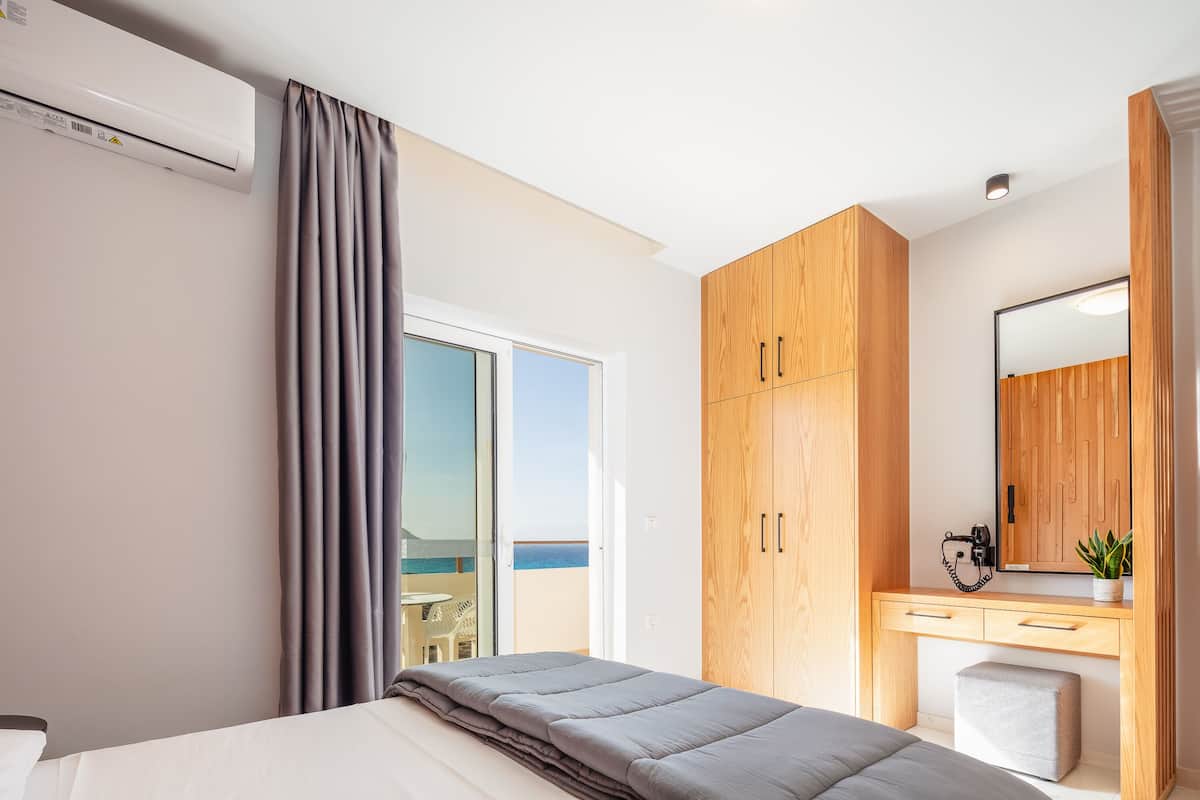 Une chambre à coucher comprenant un lit, une armoire en bois, un bureau avec un miroir et une vue sur la mer, visible à travers une porte coulissante.