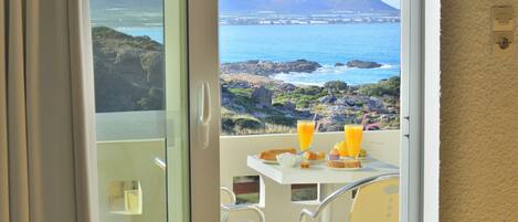 A balcony with a table set for breakfast, overlooking a coastal landscape with rocky terrain and a mountain in the distance.